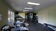 This fitness center is thoughtfully designed to accommodate a wide range of exercise needs within a single open floor layout. The space is lined with large mirrors along the walls, creating a sense of expanded space while allowing users to monitor their form during workouts. Bright rectangular overhead lighting panels, complemented by ceiling fans, ensure the room remains well-lit and comfortable during use.

The cardio section features a row of treadmills and an elliptical trainer from True Fitness, positioned along one side of the room. Adjacent to this area, a multi-station cable machine provides versatile strength training options. A dedicated free weights section includes a full dumbbell rack with a wide range of weights, supported by additional mirror panels for proper form guidance.

The flooring consists of durable rubber matting, providing both safety and comfort during high-impact activities. Natural light filters in through windows with shutters on one side, adding a pleasant ambiance to the workout environment. The overall layout is practical and spacious, offering an efficient flow between cardio, strength, and free weight areas.