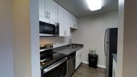 The kitchen is thoughtfully designed to maximize functionality within its compact footprint. White shaker-style cabinetry lines the walls, extending to the ceiling to provide generous storage space, while dark countertops offer a striking contrast that anchors the room's clean aesthetic. The single-level layout arranges all key appliances along one continuous run, creating an efficient workflow. Stainless steel appliances include a built-in over-the-range microwave, an electric range with a smooth cooktop, a dishwasher, and a full-sized refrigerator, all contributing to a cohesive, modern look. A double-basin stainless steel sink is centrally positioned beneath upper cabinets, making it easily accessible. The wood-look flooring adds warmth to the otherwise neutral palette of whites and grays. Recessed flush ceiling lighting provides even illumination throughout the space. Clean white baseboards and smooth walls complete the polished, move-in-ready appearance. Overall, the kitchen balances practicality with a contemporary design sensibility, offering all essential amenities in a streamlined, well-organized layout.