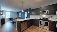 This thoughtfully designed single-floor residence combines modern aesthetics with practical living. The open-concept layout connects the kitchen and living areas seamlessly, anchored by bold teal and steel-blue accent walls that lend the space a distinct, contemporary character throughout.

The kitchen is a standout feature, showcasing dark wood-grain cabinetry with sleek bar-pull hardware, light quartz countertops, and a classic white penny-tile backsplash. Stainless steel appliances — including a gas range, over-the-range microwave, dishwasher, and refrigerator — complement the design. A peninsula with seating provides an inviting casual dining option, while pendant lighting overhead adds warmth and style.

The living area is generous and comfortable, with rich hardwood-style flooring, a large area rug, and access to an outdoor balcony through a glass door that fills the room with natural light and outdoor views.

The bedroom is a serene retreat finished in soft blue-grey tones, with plush carpeting, large windows that invite abundant natural light, and ample space for furnishings. A ceiling fan adds everyday comfort.

The bathroom is clean and refined, featuring a full soaking tub and shower enclosed by floor-to-ceiling white subway tile with polished chrome fixtures — a timeless and elegant finish that rounds out this well-balanced residence.