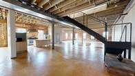 Located at 1220 Harrison Ave in Cincinnati, OH, this striking industrial loft conversion blends raw architectural character with functional living across two floors. The main level commands attention with its vast, open layout, showcasing polished amber-stained concrete floors, exposed wooden beam ceilings, and structural columns that pay homage to the building's industrial heritage. A bold black steel staircase anchors the open living area, leading to the upper level, while a kitchen with stainless steel appliances and butcher block countertops provides a modern counterpoint to the surrounding industrial aesthetic.

Throughout the space, original exposed brick walls add warmth and texture, particularly in the bedrooms and common areas, where natural light pours in through large windows dressed with sheer curtains. The primary bedroom offers generous proportions, complete with brick accent walls and soft carpeting. Three full bathrooms serve the residence, including one featuring stone tile surrounding a tub-shower combination. A dedicated laundry room houses a full-size washer and dryer. Four closets and two hallways ensure ample storage and connectivity throughout. The upper mezzanine level provides additional flexible space with panoramic windows offering views of the surrounding Cincinnati skyline.