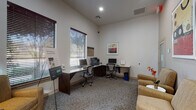 This single-floor business center is thoughtfully designed to accommodate both productive work and relaxed waiting. The space is divided into two functional zones that blend seamlessly together. The workstation area features two dedicated desks arranged in an L-shaped configuration, each equipped with a desktop computer and ergonomic task chair. A multifunction printer is conveniently positioned nearby, making the space fully operational for everyday business needs. Patterned carpeting runs throughout, adding texture and warmth to the neutral, light-toned walls.

The lounge area offers comfortable upholstered armchairs arranged around a small round side table, creating an inviting spot to relax or wait. Decorative framed artwork adorns the walls, contributing to a polished and welcoming ambiance. Tall ceilings lend an open, airy feel to the room, while horizontal blinds on the windows allow for controlled natural light.

A set of glass-paned double doors connects the business center to an adjacent common area, enhancing the sense of openness and providing visual connectivity to the broader facility. A secondary exterior door offers direct outdoor access. The overall design balances functionality with comfort, making this space suitable for residents or visitors seeking a convenient, professional environment.