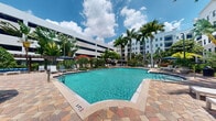 This inviting outdoor amenity space centers around a generously sized swimming pool with crystal-clear turquoise water, framed by an elegant paver deck in a warm, multi-toned stone pattern. The pool area exudes a resort-like ambiance, with the deck providing ample space for lounge chairs and sunbeds positioned to take full advantage of the open sky above. Tall royal palm trees line the perimeter, lending a distinctly tropical character to the setting, while carefully manicured shrubs and tropical plantings add lush greenery throughout. Shade umbrellas are thoughtfully placed around the deck, offering relief from the sun during warm days. The surrounding multi-story residential buildings create a sense of enclosure and privacy, forming a sheltered courtyard atmosphere. The architectural style of the buildings features clean lines with balconies overlooking the pool below. The combination of vibrant blue water, warm paver tones, and verdant landscaping creates a bright, welcoming environment ideal for relaxation and recreation. Whether lounging poolside or taking a swim, this outdoor space delivers a polished, amenity-rich experience reminiscent of a upscale resort setting.
