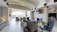 This elegant single-floor lounge space showcases a refined and welcoming atmosphere through thoughtful architectural design and upscale furnishings. The room is defined by its soaring ceilings accented with curved archways and decorative beams, lending a grand yet comfortable feel to the interior. Large, floor-to-ceiling arched windows at the far end of the space invite an abundance of natural light while offering views of a lush outdoor area, seamlessly blending the interior with the exterior environment. The flooring throughout is composed of large-format light-toned tiles, contributing to the space's open and airy ambiance. A centerpiece fireplace clad in clean, light stone tiles anchors one wall, accompanied by a wall-mounted flat-screen display above, creating a focal point for relaxation and gathering. The seating arrangement features a mix of neutral-toned sofas, accent chairs, and side tables arranged on a patterned area rug, offering multiple comfortable conversation zones. Decorative elements such as floral arrangements, sculptural accents, and carefully placed greenery add warmth and visual interest throughout. The overall design palette of soft whites, warm beiges, and muted blues creates a cohesive, sophisticated aesthetic suited for communal lounging and social interaction.