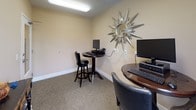 Located at 4085 Warner Ave in Landside Hills, Maryland, this single-floor business center offers a practical and well-appointed workspace designed for productivity. The space is divided into distinct workstation areas, each equipped with desktop computers, keyboards, and monitors, accommodating multiple users simultaneously. Seating options include both standard office chairs and swivel bar-height stools paired with a tall round table, providing flexibility for different working preferences.

The interior features neutral beige walls complemented by a warm terracotta accent wall, creating a welcoming yet focused atmosphere. Carpet flooring runs throughout, adding a layer of comfort underfoot. Overhead fluorescent lighting ensures the space remains well-lit, while a narrow window on the accent wall allows a touch of natural light to filter in, offering a view of the surrounding building exterior.

A decorative starburst mirror adorns one wall, adding a stylish accent to the otherwise utilitarian environment. A dark-finished console table with carved detailing and decorative ceramic vessels serves as an additional surface and subtle design element. The room is accessed through a glass-paneled door, contributing to an open and accessible feel. Overall, this business center is thoughtfully arranged to support computing needs in a tidy, comfortable, and professional setting.