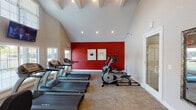 This single-story fitness center is organized into two interconnected zones, linked by a tiled entryway and glass-paneled French doors that allow natural light to flow throughout. The architecture is defined by soaring vaulted ceilings with exposed beam detailing and recessed lighting, creating an open, airy atmosphere across both sections.

The cardio-focused room features multiple treadmills and elliptical machines positioned to take advantage of large, blinds-fitted windows that overlook the surrounding outdoor area. A bold red accent wall serves as a striking focal point, complemented by framed wall art and a wall-mounted television for entertainment during workouts. Carpet tile flooring in a neutral tone provides comfort underfoot throughout both areas.

The second room is dedicated to group and functional training, housing a row of red spinning bikes, stability balls in assorted sizes, medicine balls on a wall-mounted rack, resistance equipment, and a water cooler for hydration. A full-length decorative mirror and additional windows enhance the sense of space. Framed informational signage is posted near the entrance, and the overall color palette of warm neutrals with crisp white trim creates a clean, motivating environment well-suited for a range of fitness activities.