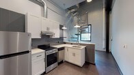 This two-story loft blends industrial character with modern finishes throughout its thoughtfully designed layout. The main level centers around a well-appointed kitchen featuring white shaker cabinetry, light countertops, a gray tile backsplash, and stainless steel appliances including a range and dishwasher. Exposed HVAC ductwork runs overhead, complementing the raw industrial aesthetic, while pendant lights provide focused task lighting. Polished concrete floors extend across the main level, adding to the urban loft atmosphere. A peninsula with a built-in sink separates the kitchen from the open living area, which benefits from soaring ceilings and exposed brick walls. A sculptural metallic spiral staircase serves as both a functional and architectural centerpiece, connecting the main level to the upper floor. The upper level houses a bedroom characterized by dramatic exposed steel trusses and dark wood ceiling beams, paired with warm hardwood-style flooring for contrast. The bathroom features large-format neutral tile flooring, built-in wall niches for storage, and a clean, minimalist design. A window on the main level draws in natural light, enhancing the open and airy quality of this distinctive industrial-inspired space.