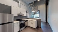 This two-story loft blends industrial character with modern finishes throughout its thoughtfully designed layout. The main level centers around a well-appointed kitchen featuring white shaker cabinetry, light countertops, a gray tile backsplash, and stainless steel appliances including a range and dishwasher. Exposed HVAC ductwork runs overhead, complementing the raw industrial aesthetic, while pendant lights provide focused task lighting. Polished concrete floors extend across the main level, adding to the urban loft atmosphere. A peninsula with a built-in sink separates the kitchen from the open living area, which benefits from soaring ceilings and exposed brick walls. A sculptural metallic spiral staircase serves as both a functional and architectural centerpiece, connecting the main level to the upper floor. The upper level houses a bedroom characterized by dramatic exposed steel trusses and dark wood ceiling beams, paired with warm hardwood-style flooring for contrast. The bathroom features large-format neutral tile flooring, built-in wall niches for storage, and a clean, minimalist design. A window on the main level draws in natural light, enhancing the open and airy quality of this distinctive industrial-inspired space.