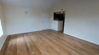 This single-floor space offers a clean, functional layout centered around an open living area. The room is finished with wide-plank hardwood flooring in warm honey tones that run throughout, creating a cohesive and inviting atmosphere. The walls are painted in a neutral off-white, providing a bright and airy feel that makes the space feel open and versatile.

A standout feature is the pass-through kitchen, which opens directly into the living area via a half-wall counter. The kitchen is equipped with dark cabinetry and recessed ceiling lighting, offering a striking contrast against the lighter tones of the main living space. This open-concept design between the kitchen and living area promotes an easy flow and connectivity throughout the unit.

The entry door is positioned along one wall of the living area, offering direct access into the main space. A wall-mounted light fixture near the entry adds a subtle decorative touch. The ceiling features a centrally placed vent, indicating a climate control system integrated into the structure. The overall design is straightforward and practical, with an emphasis on clean lines, durable materials, and an open feel that makes the space adaptable to a variety of uses and furnishing styles.