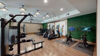 This thoughtfully designed fitness center offers a generous, open-plan exercise space complemented by a dedicated entrance area. The main workout floor is finished with warm hardwood-style flooring throughout, creating an inviting and clean aesthetic that sets the tone for the entire facility.

The exercise room is outfitted with an impressive array of equipment to support a full range of training goals. Strength training options include a multi-functional cable and pulley machine, weight racks loaded with free weights and barbells, adjustable benches, and a dedicated squat and lifting station. For cardio, the space features multiple treadmills, stair climbers, and stationary bikes, all thoughtfully arranged to allow ample room between machines.

Distinctive design elements elevate the space beyond a typical gym. A lush vertical green wall adds a striking natural accent, while a slatted wood feature wall introduces warmth and texture. Bold pendant lighting hangs from the ceiling in a clustered arrangement, complemented by ceiling fans that ensure comfortable airflow during workouts. Large windows along one side of the room invite natural light and provide views of the outdoors. A large display screen mounted on one wall adds a modern, motivating touch. Mirrors are strategically placed to expand the sense of space and support proper form during training sessions.