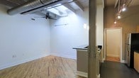 This single-floor space showcases a compelling blend of industrial and modern design. The open-plan layout is anchored by a prominent structural steel column with riveted detailing, a hallmark of industrial loft architecture that adds both character and visual interest. Hardwood floors in a warm, natural tone run throughout, providing a contrast to the industrial overhead elements.

The ceiling features exposed ductwork and wooden slat detailing, contributing to the loft's raw aesthetic. A ceiling fan and track lighting system illuminate the main living area, while pendant lights hang near the kitchen bar, adding a refined touch. The kitchen area includes a dark-topped bar counter, offering a functional space for casual dining or food preparation, with additional cabinetry visible beyond.

Tall walls painted in a clean, neutral white maximize the sense of height and openness, while allowing flexibility for personalization. A doorway visible at the rear hints at additional rooms or utility spaces. Overall, the space balances an airy, expansive feel with distinct industrial character, making it a versatile and visually dynamic environment.
