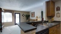 This single-floor space is thoughtfully laid out to maximize functionality and comfort. The kitchen is a standout feature, equipped with dark granite-style countertops that extend into a peninsula, offering additional prep space and separating the kitchen from the living area. A stainless steel sink and wooden cabinetry complete the kitchen's practical design. The open-concept layout allows the kitchen to flow seamlessly into the living room, which is bright and welcoming thanks to a glass-paneled entry door that draws in natural light. Neutral-toned walls and carpet throughout contribute to a cohesive, understated aesthetic.

A dedicated dining area is situated adjacent to the living space, accommodating a round table and chairs, with warm overhead lighting setting an inviting tone. A carpeted hallway connects the main living areas to the private rooms beyond, lined with multiple doors that suggest ample storage and bedroom access. Ceiling-mounted lighting illuminates the corridor effectively. Textured ceilings are consistent throughout the space, and decorative touches such as framed artwork and potted greenery enhance the overall warmth and livability of the home.