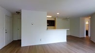 This single-floor space offers a clean, open layout designed for comfortable everyday living. The main living area features light-toned hardwood-style flooring that runs throughout, contributing to a bright and cohesive aesthetic. Neutral white walls provide a versatile backdrop, allowing for a variety of interior design choices.

The kitchen is partially open to the living area via a pass-through counter, offering both visual connectivity and practical functionality. Dark wood cabinetry provides ample storage and creates a subtle contrast against the lighter tones of the surrounding space. Recessed lighting above the kitchen ensures the area is well-illuminated for meal preparation and daily tasks.

A ceiling fan is installed in the adjacent area, supporting air circulation and adding a utilitarian element to the space. The overall design emphasizes an open flow between the kitchen and living zones, making the space feel expansive and easy to navigate. Clean lines, minimal ornamentation, and a neutral color palette give this space a move-in-ready quality that appeals to a wide range of occupants. The layout is straightforward and efficient, making it well-suited for both individuals and small households seeking a functional, low-maintenance living environment.