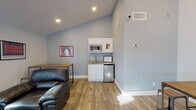 Located at 1300 E Fort Lowell Rd in Tucson, Arizona, this single-floor community lounge space is designed with a strong University of Arizona Wildcats theme throughout. The interior features light blue walls, wood-look flooring, and soaring vaulted ceilings that lend an open, airy feel to the space.

The main lounge area is furnished with a full black leather sectional sofa, a matching armchair, and a tufted ottoman, all arranged to encourage social gathering. Large floor-to-ceiling windows along one wall flood the space with natural light while offering views of mature trees and the surrounding outdoor environment.

A dedicated dining and study area features industrial-style high-top tables paired with metal and wood stools, complemented by a wall-mounted flat-screen TV — ideal for group viewing or casual gatherings. University of Arizona artwork, including "Bear Down" and Wildcats mascot prints, decorates the walls throughout, reinforcing the spirited collegiate atmosphere.

A compact kitchenette serves the space, equipped with white cabinetry, a microwave, a small sink with stone-look countertop, and a stainless steel mini-refrigerator — providing convenient amenities for residents or guests. Recessed lighting and natural light combine to keep the space bright and welcoming. The overall design balances comfort, functionality, and school pride in a clean, modern setting.