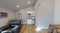 Located at 1300 E Fort Lowell Rd in Tucson, Arizona, this single-floor community lounge space is designed with a strong University of Arizona Wildcats theme throughout. The interior features light blue walls, wood-look flooring, and soaring vaulted ceilings that lend an open, airy feel to the space.

The main lounge area is furnished with a full black leather sectional sofa, a matching armchair, and a tufted ottoman, all arranged to encourage social gathering. Large floor-to-ceiling windows along one wall flood the space with natural light while offering views of mature trees and the surrounding outdoor environment.

A dedicated dining and study area features industrial-style high-top tables paired with metal and wood stools, complemented by a wall-mounted flat-screen TV — ideal for group viewing or casual gatherings. University of Arizona artwork, including "Bear Down" and Wildcats mascot prints, decorates the walls throughout, reinforcing the spirited collegiate atmosphere.

A compact kitchenette serves the space, equipped with white cabinetry, a microwave, a small sink with stone-look countertop, and a stainless steel mini-refrigerator — providing convenient amenities for residents or guests. Recessed lighting and natural light combine to keep the space bright and welcoming. The overall design balances comfort, functionality, and school pride in a clean, modern setting.