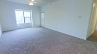 This single-floor space offers a clean, open layout designed to maximize comfort and functionality. The main living area is generously proportioned, providing ample room for a variety of furniture arrangements and lifestyle needs. Soft, plush wall-to-wall carpeting covers the floors, adding warmth and comfort underfoot throughout the space. The room is bathed in natural light, courtesy of multiple windows — including a notably large multi-pane window that draws in bright, ambient daylight and creates an airy, welcoming atmosphere. A ceiling fan is centrally mounted overhead, ensuring efficient air circulation year-round. The walls are finished in a neutral, light tone that enhances the sense of openness and pairs well with a wide range of décor styles. A glass-panel interior door adds a subtle architectural detail, allowing light to flow between spaces while maintaining a sense of separation. The overall ambiance is calm and understated, offering a bright, neutral canvas that is move-in ready and easily personalized. This is a practical and comfortable space that balances simplicity with livability on a single, accessible level.