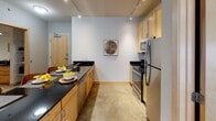 This well-appointed single-floor residence offers a thoughtfully designed open-concept layout that seamlessly connects the kitchen, dining, and living areas. The kitchen is outfitted with stainless steel appliances, light maple cabinetry, dark granite countertops, a built-in dishwasher, and a breakfast bar with seating — ideal for both meal prep and casual dining. Track lighting overhead enhances the clean, modern aesthetic. The living area provides a comfortable and stylish retreat, featuring large windows with roller shades that allow natural light to fill the room, complemented by views of the surrounding urban environment. A consistent grey, white, and yellow color palette ties the space together with a modern, cohesive feel. The primary bedroom includes large windows, ample floor space, and a dedicated desk area, while the second bedroom comfortably accommodates two twin beds with built-in wall shelving for added storage. Two bathrooms serve the unit — one featuring a dark granite vanity with maple cabinetry and a stacked washer and dryer conveniently integrated into the space, and the other a compact full bath with tile flooring and a walk-in closet area. Throughout the unit, polished concrete flooring, neutral wall tones, and contemporary artwork contribute to a refined, urban living experience.