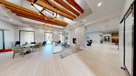 Located at 9550 W Sahara Ave in Las Vegas, this generously sized single-floor community clubhouse delivers an impressive blend of modern design and welcoming comfort. The grand main living area immediately captures attention with its soaring ceilings adorned by rich, exposed wood beams and a striking geometric chandelier that anchors the space. Light wide-plank hardwood flooring flows throughout, complementing the crisp white walls and abundant natural light streaming through large black-framed glass doors and windows. A sleek double-sided fireplace serves as a dramatic focal point, flanked by plush seating areas that invite relaxed conversation. A separate lounge zone features a wall-mounted TV, additional fireplace, and direct access to the outdoor courtyard through floor-to-ceiling glass doors. The recreation room houses a full-size billiards table alongside built-in banquette seating and views of mature palms outside. Stylish barn doors with frosted glass panels lend a contemporary touch to interior transitions. A dedicated service counter with warm wood cabinetry offers a convenient refreshment station. Two well-appointed restrooms feature trough sinks and clean tile finishes. The space also includes a casual gaming area, rounding out a versatile and thoughtfully designed facility.