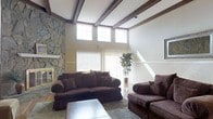 The living area of this single-story space makes a striking impression with its soaring vaulted ceiling adorned with rich, dark-stained exposed wood beams that add warmth and architectural character. A row of clerestory windows positioned high along the upper wall floods the room with natural light, creating a bright and airy atmosphere throughout the day. A floor-to-ceiling stacked stone fireplace anchors one wall, serving as a bold focal point that adds a rustic yet refined aesthetic to the space. The walls are finished in a neutral tone, complemented by classic wainscoting details that lend a traditional elegance to the room. Sliding glass doors and additional windows provide further natural light and potential access to an outdoor area. The great room comfortably accommodates generous seating arrangements, as evidenced by the plush, dark upholstered sofas arranged around a tiled coffee table atop a soft area rug. The combination of stone, wood, and neutral finishes creates a cohesive, inviting ambiance that is both casual and sophisticated, making this living space well-suited for both everyday living and entertaining.