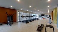 This single-level fitness facility offers a clean, contemporary design built to support a comprehensive workout experience. The main exercise area is generously proportioned, featuring polished concrete flooring that lends an industrial yet refined aesthetic throughout the space. A striking terracotta-red accent wall near the entrance adds a bold visual element, contrasting effectively with the otherwise neutral white walls and ceiling.

Natural light flows in through a wide span of louvered floor-to-ceiling windows along one wall, creating a bright and energizing atmosphere while maintaining privacy and airflow control. Recessed ceiling lighting supplements the natural light, ensuring the space remains well-illuminated at all times.

The cardio zone is well-stocked with a long row of treadmills and stair climbers positioned along the windowed wall, alongside rowing machines on the open floor. Additional strength and cable equipment can be seen toward the perimeter, complemented by a wall-mounted television for entertainment. A mirrored wall enhances the sense of depth and space, making the facility feel expansive and open.