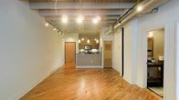 This single-floor urban loft showcases a compelling blend of industrial design and modern living. The open-plan layout creates a generous, flowing living area anchored by warm hardwood floors that run throughout the space, lending a sense of continuity and natural warmth.

The kitchen sits at the heart of the space, defined by a sleek peninsula-style island with a dark countertop. Pendant lighting hangs above the island, casting focused illumination over the prep and dining area, while the kitchen is equipped with stainless steel appliances including a microwave and range. Cabinetry in a warm wood finish ties the kitchen to the broader aesthetic of the space.

Overhead, the industrial character of the loft is unmistakable. Exposed ductwork, structural beams, and a concrete ceiling are left intentionally visible, creating an authentic urban loft atmosphere. Track lighting and pendant fixtures are thoughtfully distributed to ensure the open floor plan is well-lit throughout.

Two interior doors — one in a natural wood finish and another featuring a louvered panel design — provide access to additional rooms or storage areas, hinting at further functional spaces beyond the main living area. The combination of raw architectural elements and warm finishes makes this loft a distinctive and characterful space suited to modern urban living.