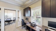 Located at 900 Grand Central Dr in Hamilton, NJ, this professional suite offers a polished and functional environment designed to support a variety of business needs. The space is thoughtfully divided into two distinct areas: a private office and a conference room, connected through an elegantly framed doorway with a dark-trimmed glass door that allows natural light to flow between rooms.

The office area features built-in dark espresso cabinetry with overhead storage and a granite-topped L-shaped workstation, providing ample workspace. A large window with slatted blinds offers views of lush greenery, keeping the workspace bright and inviting. The tray ceiling with crown molding adds a refined architectural touch throughout.

The conference room is equally impressive, centered around a sleek oval dark table surrounded by ergonomic task chairs. Two generously sized windows flood the room with natural light, while a stylish multi-light pendant chandelier hangs above. A built-in whiteboard panel offers practical presentation capabilities. The tray ceiling with dark wood beam accents mirrors the sophisticated design seen throughout the suite.

Warm neutral tones, crown molding, and carpeted flooring unify the space, creating a professional yet welcoming atmosphere well-suited for meetings, focused work, or client-facing interactions.