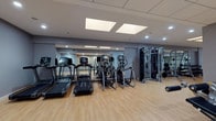 This fitness center is a comprehensively equipped facility designed to support a full range of workout routines. The space is finished with warm light wood flooring throughout, set against cool gray walls that create a clean, focused atmosphere. Recessed lighting and skylights provide bright, even illumination across the entire floor.

The cardio zone features multiple treadmills and elliptical trainers, each equipped with individual display screens for entertainment and performance tracking. Full-length mirrors line the walls, enhancing the sense of openness and allowing users to monitor their form during exercise.

The strength training area is generously appointed with a power rack, a multi-station cable machine, a full dumbbell rack with a variety of weights, adjustable benches, and several selectorized machines targeting different muscle groups. TRX suspension trainers and a stationary cycling bike add functional and low-impact training options.

A glass-partitioned area is visible at one end of the facility, providing a designated enclosed zone with patterned carpet flooring — suitable for stretching, group classes, or separate training use. The overall layout is thoughtfully organized to allow for comfortable movement between equipment stations, making this fitness center a well-rounded and functional space for users of all fitness levels.