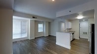 Located at 101 Summer Street in Stamford, Connecticut, this single-floor residence offers a clean, contemporary design with an open-concept layout that seamlessly connects the living and kitchen areas. The kitchen is outfitted with crisp white cabinetry, granite countertops, a stainless steel sink, and integrated appliances, anchored by a peninsula that serves as both a functional workspace and a natural divider between the cooking and living zones. The living area is bright and airy, benefiting from multiple windows dressed with horizontal blinds that allow natural light to filter through while maintaining privacy. Dark hardwood-style flooring runs throughout the main living space, adding warmth and contrast to the neutral gray and white color palette of the walls and ceilings. The kitchen area transitions to a lighter wood-tone floor, subtly delineating the two spaces. Recessed lighting and flush-mount ceiling fixtures provide well-distributed artificial lighting throughout. The overall ambiance is modern and move-in ready, with clean lines, neutral tones, and a functional open floor plan that maximizes the sense of space — making it well-suited for a variety of lifestyle needs.
