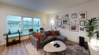 Located at 24615 64th Ave S, Unit C219 in Kent, Washington, this contemporary single-floor residence offers a well-designed layout with modern finishes throughout. Light wood-tone flooring flows seamlessly from the entry hallway through the main living areas, creating a cohesive and inviting atmosphere.

The living area is a standout space, anchored by large windows fitted with horizontal blinds that flood the room with natural light while offering views of the surrounding neighborhood. The layout comfortably accommodates a sectional sofa, a bistro-style high-top table and chairs positioned by the windows, and a gallery wall arrangement that adds personality and visual interest.

The kitchen is outfitted with contemporary two-tone cabinetry, stainless steel appliances including a refrigerator, electric range, and over-the-range microwave, complemented by a white subway tile backsplash and solid-surface countertops. A stainless steel sink with a modern faucet completes this functional and attractive cooking space.

The hallway connects the living areas to the private spaces, including a full bathroom finished with wood-grain cabinetry, a combined tub and shower, and clean white walls. Recessed lighting throughout the unit ensures well-lit spaces at every turn. The overall design reflects a clean, modern aesthetic suited for comfortable everyday living.