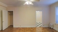 This single-floor space displays a thoughtful blend of classic architectural elements and timeless design details. The main room is finished with warm parquet wood flooring laid in a traditional pattern, adding texture and character underfoot. Crisp white walls are accented by decorative panel molding, lending a refined, formal quality to the space. Crown molding runs along the ceiling perimeter, reinforcing the classic aesthetic throughout. A ceiling fan with a built-in light fixture is centrally mounted, providing both airflow and ambient lighting.

An open doorway connects the main room to an adjoining space, visible through a clean white-trimmed threshold. This secondary room features a striking black-and-white checkered tile floor, creating a bold visual contrast to the warmer tones of the primary space. A small chandelier is partially visible within this adjoining room, hinting at an elevated level of detail carried throughout the unit.

The overall ambiance is clean and neutral, with the white walls and bright lighting creating an airy, open feel. The combination of parquet wood, decorative molding, and checkered tile gives the space a distinctive character rooted in classic residential design.