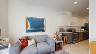 This thoughtfully designed single-floor residence showcases a clean, modern aesthetic with white walls and light wood-style flooring that flows seamlessly throughout the space. The open-concept living and kitchen area forms the heart of the unit, where a comfortable lounge space anchored by a gray loveseat and bold abstract artwork transitions naturally into a fully equipped kitchen. The kitchen features stainless steel appliances, including a range, dishwasher, and over-the-range microwave, complemented by two-tone cabinetry — crisp white upper cabinets paired with wood-toned lower cabinets — and a classic white subway tile backsplash. A breakfast bar with wood stools provides a casual dining option. The living area benefits from large windows that invite generous natural light and offer views of the surrounding urban streetscape. The bedroom is a calm retreat, featuring a sliding closet door, warm bedside lighting, and curated artwork above the bed, creating an inviting and restful atmosphere. The bathroom maintains the unit's contemporary style with wood-toned cabinetry, a large mirror, and clean white walls. An in-unit washer and dryer add a practical convenience. Throughout, the space balances functional urban living with a warm, curated design sensibility.