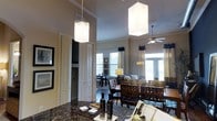 This thoughtfully designed single-floor residence showcases an open-concept layout that seamlessly connects the kitchen, dining, and living areas into one cohesive, flowing space. The kitchen features a striking dark granite countertop and is anchored by two large rectangular pendant lights that hang dramatically from the soaring ceilings, immediately drawing the eye upward and emphasizing the impressive vertical scale of the interior. Crown molding adds a refined architectural touch throughout.

The dining area is furnished with rich, dark wood chairs with woven leather seats, lending a sophisticated and warm character to the space. Adjacent to the dining area, the living room is generously proportioned and bathed in natural light streaming through multiple large windows framed by navy and white curtains, creating a bright and airy atmosphere. A ceiling fan with integrated lighting serves the living area, ensuring comfort year-round.

French doors along the far wall provide direct access to the outdoors, further enhancing the sense of openness and bringing in additional natural light. Neutral warm-toned walls complement the dark furnishings throughout, creating a balanced and inviting ambiance. A hallway with closet access provides practical utility without disrupting the open feel of the main living spaces. Overall, this residence combines elegant design details with functional living areas.