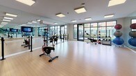 This single-floor yoga and fitness studio is designed with both functionality and aesthetics in mind. The space features a generously sized open floor plan finished with light hardwood flooring that creates a clean, welcoming atmosphere throughout. Large floor-to-ceiling windows line one wall, flooding the studio with abundant natural light and offering views of the surrounding neighborhood, enhancing the sense of openness and connection to the outdoors. Full-length wall mirrors are installed along multiple walls, providing visual depth and supporting proper form during workouts and movement classes. Recessed panel lighting across the ceiling ensures bright, even illumination throughout the day and evening. The studio is equipped with stationary cycling bikes, stability balls of varying sizes on dedicated racks, kettlebells, a ballet barre, resistance equipment, and additional fitness accessories neatly organized along the perimeter. A large flat-screen display is mounted on one wall, supporting on-demand fitness programming. Glass-paneled doors provide access to adjacent spaces, contributing to the studio's modern, transparent aesthetic. A wall-mounted air conditioning unit ensures a comfortable environment during intense sessions. The overall ambiance is bright, clean, and professionally appointed, making it a versatile space suited for yoga, cycling, barre, and a variety of group fitness activities.
