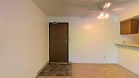 This single-floor space offers a straightforward and functional layout designed for comfortable everyday living. The entryway is finished with dark tile flooring, providing a durable and easy-to-clean transition zone as you step through the solid wood-toned front door, which is equipped with a security peephole and door closer for added safety and convenience. Moving further inside, the flooring transitions to carpet, lending a warmer, softer feel to the main living area. The walls are finished in a clean, neutral white, creating a bright and open atmosphere that serves as a versatile backdrop for a variety of interior styles. A ceiling fan with an integrated multi-bulb light fixture is centrally positioned overhead, offering both air circulation and ambient lighting throughout the space. The overall ambiance is clean and understated, with a practical design that emphasizes ease of use and adaptability. The simple, uncluttered interior presents an excellent opportunity for personalization and customization to suit a wide range of needs and preferences.