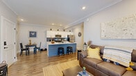This well-appointed single-floor residence showcases a thoughtfully designed open-concept layout that seamlessly connects the kitchen, dining, and living areas. The kitchen is a standout feature, boasting white shaker cabinetry, a striking blue shiplap island with a built-in sink and bar seating for two, stainless steel appliances, and a navy blue tile backsplash that adds a bold contrast to the crisp white countertops. Recessed lighting and crown molding throughout lend a polished, cohesive aesthetic to the main living spaces. The living room offers comfortable proportions, a wall-mounted TV, and access to a private exterior door, filling the space with natural light. A separate dining nook provides an intimate setting for meals. The primary bedroom is generously sized, featuring multiple windows dressed with neutral linen curtains that invite abundant natural light, and connects to a well-appointed bathroom with white shaker vanity cabinetry, black hardware accents, and hardwood-style flooring. A second bedroom mirrors the serene, light-filled character of the primary suite. Warm hardwood-style flooring flows consistently throughout the entire residence, unifying the space with a modern yet inviting character.