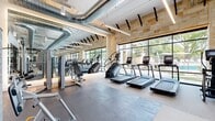 This expansive fitness center offers a thoughtfully designed, single-floor exercise space that blends modern industrial aesthetics with upscale finishes. The open floor plan is divided into distinct functional zones, seamlessly flowing from one area to the next. Exposed ductwork and sleek linear lighting run overhead, complementing the high ceilings that lend the space a loft-like openness.

Natural limestone accent walls add warmth and texture, contrasting beautifully with polished concrete-style flooring and light-toned carpet tiles that define different workout areas. Floor-to-ceiling glass windows and doors line one side of the facility, flooding the interior with abundant natural light and offering views of an outdoor pool and lush greenery beyond.

The cardio zone is equipped with a full lineup of treadmills, ellipticals, stair climbers, stationary bikes, and rowing machines, all arranged to face the panoramic windows. The strength training area features a comprehensive selection of Matrix cable and selectorized machines, flanked by floor-to-ceiling mirrors with integrated backlighting. A dedicated free weights zone includes a full dumbbell rack and functional training rigs. A large open stretch and movement area with mirrored walls completes the facility, providing ample space for group exercise or individual training sessions.