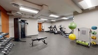This fitness center offers a comprehensive workout environment designed to accommodate a wide range of exercise routines. The space is laid out across a single floor and is organized to maximize functionality, with clearly defined zones for cardio, strength training, and flexibility work.

The cardio section is equipped with multiple treadmills, an elliptical cross-trainer, and a stationary exercise bike, each featuring integrated digital displays for tracking workout metrics. A rowing machine rounds out the cardio offerings. The strength training area includes a multi-station cable machine and a dedicated free weights section, featuring a full rack of dumbbells displayed on a tiered stand, accompanied by an adjustable workout bench positioned in front of a large wall-mounted mirror. The mirror expands the visual space and allows users to monitor their form during exercises.

A flexibility and functional fitness area is stocked with stability balls in various sizes and a yoga mat, providing ample space for stretching and bodyweight exercises. The flooring throughout is a durable, speckled rubber surface well-suited for heavy equipment and high-traffic use.

The interior design features neutral gray and white walls accented by a bold orange feature wall, adding energy and visual interest to the space. Overhead fluorescent panel lighting ensures the entire area is brightly and evenly illuminated. A water fountain and restroom access are conveniently located within the facility.