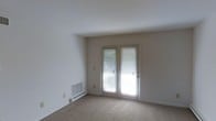 This single-floor residential space offers a clean, understated interior defined by neutral tones and practical finishes throughout. The living area is finished with wall-to-wall carpeting that adds a sense of warmth underfoot, while the bright white walls and ceilings create an open, airy atmosphere. A standout feature of the room is the set of French doors, which allow generous natural light to fill the space and provide a view of the greenery outside. The doors are fitted with horizontal blinds, offering privacy and light control as needed. Heating is provided by a baseboard heating unit positioned along the lower wall, ensuring efficient climate management throughout the colder months. A floor-level wall vent further supports the home's HVAC system. The room's layout is straightforward and versatile, lending itself well to a variety of furniture arrangements. The overall ambiance is calm and functional, with a simple, no-frills design that serves as an adaptable canvas for personalization. The space is well-suited for comfortable everyday living.