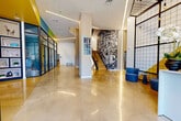 Located at 1 Green Island Boulevard in Worcester, Massachusetts, The Revington presents a thoughtfully designed ground-floor common area that makes a striking first impression. The expansive lobby showcases gleaming polished concrete floors that reflect the recessed linear lighting overhead, creating a bright and welcoming atmosphere. Bold pops of yellow, teal, and green punctuate the white walls and ceiling accents, lending the space a contemporary, energetic personality.

At the heart of the lobby stands a sleek reception desk enclosed within a dramatic black steel-and-glass grid partition, complemented by blue lounge seating and a vivid yellow geometric backdrop. A large-scale black-and-white mural lines the staircase wall, adding an artistic focal point to the open circulation area. Floor-to-ceiling windows at the entrance flood the space with natural light and offer views of the surrounding streetscape.

Glass-partitioned meeting and lounge rooms line the corridor, maintaining visual openness while providing functional, private spaces. A dedicated mail room features a vibrant teal accent wall and a full bank of resident mailboxes. Additional amenities include a coffee station with built-in display shelving. Throughout, the design balances bold aesthetics with practical functionality, creating an inviting and dynamic environment.