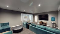 This single-floor lounge and screening space is thoughtfully designed for comfortable group entertainment and socializing. The room is finished with neutral grey walls and smooth ceilings equipped with recessed lighting, creating a clean, modern aesthetic throughout. Dark-toned carpet flooring anchors the space and complements the overall color palette.

The seating arrangement is a standout feature, comprising multiple tufted teal velvet sofas and sectionals positioned to face a large wall-mounted flat-screen television set atop a warm wood media console. Rounded leather ottomans and tufted upholstered benches are distributed throughout, adding both functional and decorative elements. Gold-accented side tables provide convenient surfaces between seating clusters.

One wall features floor-to-ceiling sheer curtains that diffuse natural light softly into the room, hinting at an adjacent outdoor area or balcony visible through the fabric. A wall-mounted split air conditioning unit ensures climate control, while framed artwork depicting monochromatic music-themed imagery adds a curated, artistic touch to one wall.

The layout accommodates multiple seating zones, making it well-suited for both intimate gatherings and larger group screenings. The combination of plush furnishings, controlled lighting, and entertainment-focused design gives the space a welcoming, upscale lounge ambiance.