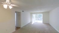 This single-floor space features a spacious, open-concept living area finished in a clean, neutral palette throughout. The living room is carpeted in a light beige tone, contributing to a warm and understated aesthetic that makes the space feel welcoming and versatile. A textured ceiling adds subtle architectural character overhead.

Natural light is a defining feature of the living area, flooding in through a large sliding glass door fitted with sheer white curtains. The door opens to a private balcony enclosed by a white railing, with views of mature greenery beyond — creating a pleasant indoor-outdoor connection. This abundant natural light enhances the airy quality of the room.

Adjacent to the living area, a doorway with a louvered bifold closet door leads to a connecting hallway or additional room, hinting at further functional spaces within the unit. The transition from carpet to hard flooring in this area suggests distinct zones for living and utility. A ceiling-mounted HVAC vent ensures year-round climate comfort. Overall, the space presents a clean, light-filled environment well-suited for comfortable everyday living.