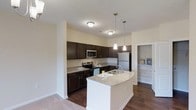 Located at 40 Stallings Mill Rd in Clayton, North Carolina, this single-floor residence offers a thoughtfully designed living space centered around a modern, open-concept kitchen. The kitchen is a standout feature, showcasing rich dark cabinetry that contrasts beautifully with light granite countertops. A curved kitchen island with an undermount sink and elegant pendant lighting serves as both a functional workspace and a natural gathering point. Stainless steel appliances, including a refrigerator, range, and over-the-range microwave, complete the well-appointed culinary space. The flooring transitions from rich dark hardwood in the kitchen and main areas to plush carpet in adjacent living spaces, adding warmth and comfort underfoot. Neutral-toned walls throughout create a clean, versatile backdrop that complements a variety of interior styles. A walk-in closet with built-in shelving is conveniently accessible from the main living area, offering excellent organizational storage. Recessed and flush-mount ceiling lighting ensures the space remains bright and welcoming throughout. Overall, the layout is practical and inviting, combining contemporary finishes with a functional design that suits modern everyday living.