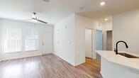 This single-floor unit showcases a clean, modern design highlighted by wood-look plank flooring that runs consistently throughout the space. The open-concept layout connects the living area to the kitchen, creating a cohesive and functional flow. Recessed lighting and ceiling fans are installed throughout, enhancing both comfort and illumination.

The kitchen is outfitted with white shaker-style cabinetry, light quartz countertops, a white subway tile backsplash, stainless steel appliances including an electric range and microwave, and a black matte faucet over a stainless undermount sink. A peninsula counter separates the kitchen from the living area, offering additional prep and seating space.

The living area is spacious and bright, with natural light entering through blinded windows near the front entry door, which also features a ceiling fan. A secondary room, accessible through a paneled interior door, benefits from two windows overlooking greenery and its own ceiling fan, making it well-suited as a bedroom or flex space.

The bathroom includes a tub-shower combination with white tile surround, a white vanity with undermount sink, chrome fixtures, and wood-look flooring. The overall palette of white walls, neutral flooring, and modern hardware gives the unit a fresh, move-in-ready appearance.