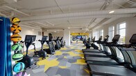 This fitness center in Savannah, Georgia, offers a comprehensive workout environment spread across a single, generously proportioned floor. The space is defined by its striking geometric carpet design in yellow and gray, lending a vibrant, energetic atmosphere throughout. White-painted ceilings with exposed ductwork and pipework give the space an industrial-modern aesthetic, while recessed and surface-mounted lighting ensures the entire floor is well illuminated.

The cardio zone is prominently featured along one side of the main floor, housing a row of TRUE-brand treadmills, upright and recumbent stationary bikes, and elliptical cross-trainers. Large windows along the exterior wall flood this section with natural light and provide open views of the surrounding Savannah landscape and balcony beyond.

A dedicated free weights area features a tiered dumbbell rack and an adjustable bench, positioned near a floor-to-ceiling mirror framed by a bold yellow accent wall. A large flat-screen TV mounted within the mirrored wall serves as a focal point for this zone. Additional functional training equipment, medicine balls, stability balls, and resistance accessories are neatly organized in a separate section. The overall ambiance is clean, modern, and motivating.