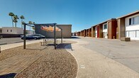 Located at 2423 W Campbell Ave in Phoenix, Arizona, this single-story residential complex offers a well-organized layout centered around a spacious open courtyard. The buildings feature a distinctive mid-century-inspired exterior with brown and white stucco facades accented by bold red entryway surrounds and matching stairwells, giving the property a recognizable and cohesive aesthetic.

The central courtyard serves as the heart of the community, providing a welcoming outdoor gathering space. A shade structure with a fabric canopy covers a picnic table and seating area, offering a comfortable retreat from the Arizona sun. Adjacent to the seating area is a standalone BBQ grill set on a paved patio, surrounded by desert-landscaped gravel that minimizes water use while maintaining a clean, low-maintenance appearance. Tall palm trees add a classic Phoenix character to the outdoor space.

On-site parking is generously available, with a dedicated paved lot providing clearly marked spaces for residents. The wide concrete pathways and circular drive throughout the property ensure easy vehicle and pedestrian access. The overall layout is open and practical, designed to accommodate the needs of its residents efficiently. The complex benefits from Phoenix's abundant sunshine and clear skies, making the outdoor amenities a functional and enjoyable extension of daily life.