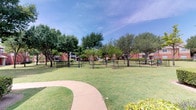 Located in Frisco, Texas, this outdoor common area serves as a central gathering and recreational space within a brick residential community. The grounds are defined by a generously sized, enclosed dog park surrounded by black iron fencing, providing a secure area for pets to roam freely. The open grassy space is complemented by areas of packed gravel and dirt, accommodating active use throughout the day.

Mature shade trees are distributed throughout the grounds, offering natural canopy cover and a sense of established landscaping. Seating benches are positioned within and around the fenced area, providing convenient spots for residents to relax while supervising their pets. A curved concrete walkway winds through the well-kept lawn, connecting different areas of the community and adding to the overall accessibility of the space.

The surrounding brick residential buildings frame the common area on multiple sides, contributing to an enclosed, neighborhood-like atmosphere. A glimpse of what appears to be a pool area is visible at the edge of the grounds, suggesting additional recreational amenities nearby. The combination of open green space, thoughtful landscaping, and dedicated pet-friendly infrastructure makes this outdoor area a functional and inviting community asset.