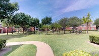 Located in Frisco, Texas, this outdoor common area serves as a central gathering and recreational space within a brick residential community. The grounds are defined by a generously sized, enclosed dog park surrounded by black iron fencing, providing a secure area for pets to roam freely. The open grassy space is complemented by areas of packed gravel and dirt, accommodating active use throughout the day.

Mature shade trees are distributed throughout the grounds, offering natural canopy cover and a sense of established landscaping. Seating benches are positioned within and around the fenced area, providing convenient spots for residents to relax while supervising their pets. A curved concrete walkway winds through the well-kept lawn, connecting different areas of the community and adding to the overall accessibility of the space.

The surrounding brick residential buildings frame the common area on multiple sides, contributing to an enclosed, neighborhood-like atmosphere. A glimpse of what appears to be a pool area is visible at the edge of the grounds, suggesting additional recreational amenities nearby. The combination of open green space, thoughtful landscaping, and dedicated pet-friendly infrastructure makes this outdoor area a functional and inviting community asset.