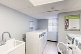 This shared laundry facility at 8950 Wyandotte St E in Windsor, Ontario, offers a clean, functional, and well-organized space designed for convenient everyday use. The room features light grey walls, a drop-ceiling with integrated lighting, and durable light wood-tone flooring that gives the space a fresh, maintained appearance.

The laundry room is equipped with multiple top-load and front-load washers and dryers by Huebsch and Coinamatic, arranged in a practical layout that maximizes the available floor space. Machines support modern cardless payment methods, offering added convenience for users. A utility sink with a white cabinet base provides additional functionality for hand-washing or pre-treating laundry.

A wall-mounted payment kiosk, a drop box for payments or requests, and clearly posted laundry instructions ensure a straightforward user experience. A fire extinguisher and smoke detector are present, reflecting attention to safety standards. A window near the entrance allows for a degree of natural light, while a grey door with a glass panel provides secure access to the room. A small seating area with a chair offers a comfortable spot to wait.

Overall, this laundry facility is a practical, tidy, and well-maintained communal space that meets the everyday laundry needs of building residents efficiently.