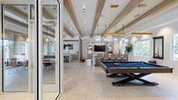 This single-story amenity space is designed in a coastal-modern style, characterized by an open, airy floor plan, large-format light-toned tile flooring, and exposed wood ceiling beams that run throughout, adding warmth and architectural interest to the predominantly white interior. Natural light flows generously through multiple windows and glass doors, reinforcing the bright, welcoming ambiance.

The main gathering area features a spacious game room equipped with a full-size pool table and a built-in entertainment wall with a wall-mounted television, open shelving, and seating for a comfortable lounge experience. Pendant lighting in seafoam glass and woven designs adds a nautical touch overhead. Adjacent to this is a wet bar area with counter seating, ideal for casual socializing.

A separate dining zone accommodates a large communal table surrounded by mid-century-style chairs in walnut and teal, illuminated by striking sculptural gold pendant lights. Banquette seating lines the windowed walls, and glass doors open directly to an outdoor patio area, seamlessly connecting interior and exterior spaces.

A private glass-enclosed office nook provides a quiet workspace, while a comfortable lounge area with a plush sofa, accent chairs, striped area rugs, and a built-in storage wall offers a refined space for relaxation. The overall atmosphere is polished yet inviting throughout.