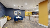 Located at 8 W King St in the heart of Lancaster, PA, this contemporary lounge offers a thoughtfully designed, single-floor space that blends professional functionality with a lively, welcoming aesthetic. The interior is anchored by a bold color palette of deep navy blue and vibrant golden yellow, carried consistently throughout the walls, furnishings, and accent elements, creating an energetic yet cohesive atmosphere.

The open main area features a variety of flexible seating arrangements, including standing-height bar tables with mid-century modern stools, intimate round tables with plush blue velvet chairs, and comfortable lounge seating with upholstered armchairs in navy and yellow. Patterned carpet tiles in neutral tones run throughout, providing texture underfoot while tying the space together. Recessed lighting and pendant fixtures with gold hardware illuminate the area evenly overhead, complemented by a suspended drop-tile ceiling.

A designated Focus Room offers a quieter retreat, its entrance marked with clear signage on a deep blue accent wall. A small utility area features a countertop with a sink, white cabinetry, and stone-look surfaces, providing practical convenience. Open shelving displays decorative accents and greenery, adding warmth throughout. Slatted wood wall panels and curated artwork further elevate the refined, modern character of this well-appointed lounge.