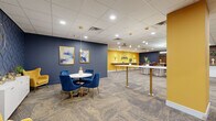 Located at 8 W King St in the heart of Lancaster, PA, this contemporary lounge offers a thoughtfully designed, single-floor space that blends professional functionality with a lively, welcoming aesthetic. The interior is anchored by a bold color palette of deep navy blue and vibrant golden yellow, carried consistently throughout the walls, furnishings, and accent elements, creating an energetic yet cohesive atmosphere.

The open main area features a variety of flexible seating arrangements, including standing-height bar tables with mid-century modern stools, intimate round tables with plush blue velvet chairs, and comfortable lounge seating with upholstered armchairs in navy and yellow. Patterned carpet tiles in neutral tones run throughout, providing texture underfoot while tying the space together. Recessed lighting and pendant fixtures with gold hardware illuminate the area evenly overhead, complemented by a suspended drop-tile ceiling.

A designated Focus Room offers a quieter retreat, its entrance marked with clear signage on a deep blue accent wall. A small utility area features a countertop with a sink, white cabinetry, and stone-look surfaces, providing practical convenience. Open shelving displays decorative accents and greenery, adding warmth throughout. Slatted wood wall panels and curated artwork further elevate the refined, modern character of this well-appointed lounge.